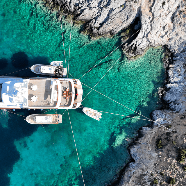 Aerial view of a yacht anchored in clear turquoise waters between rocky cliffs.